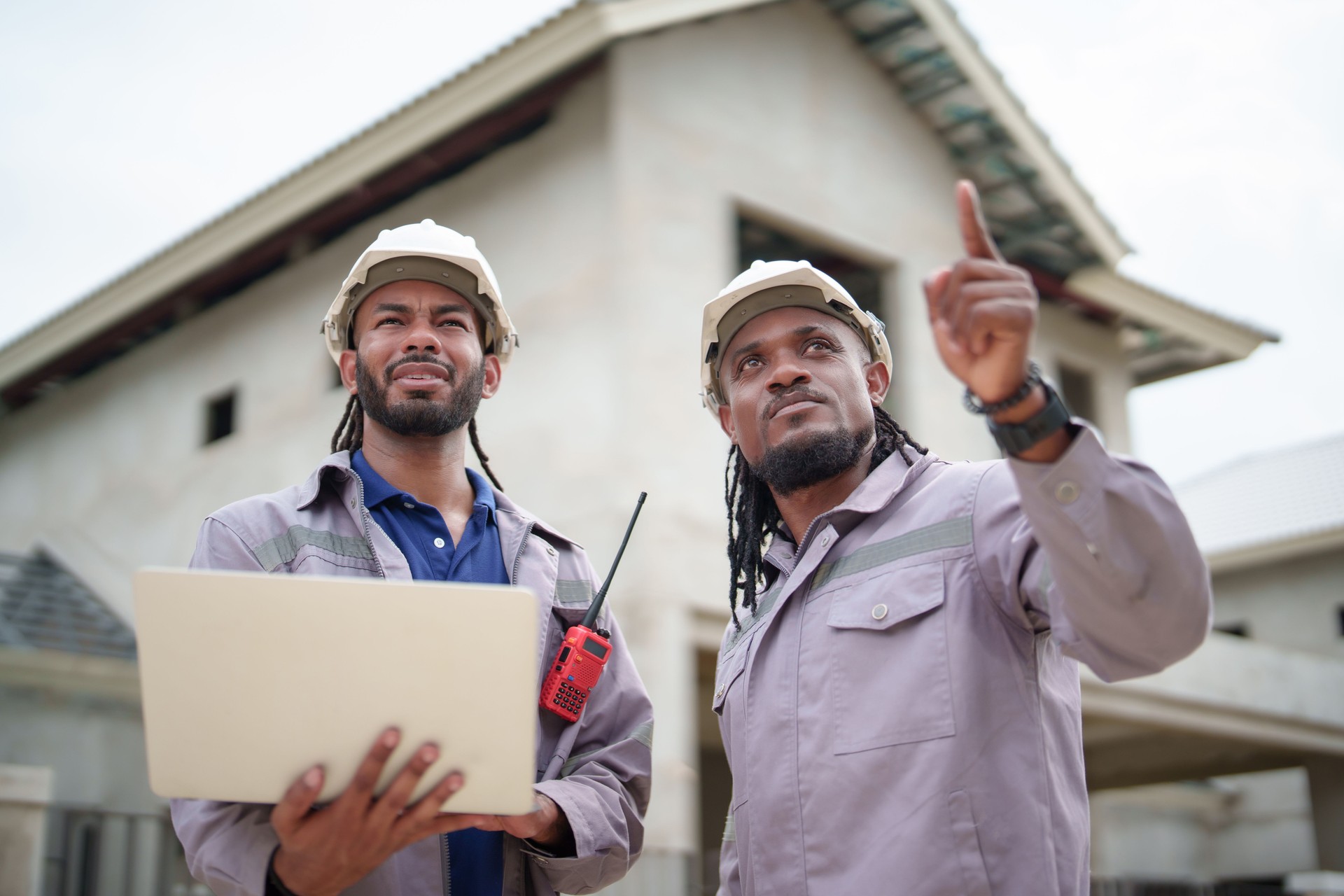 Two construction workers wearing hard hats are discussing building project. One holds laptop and other points towards construction site, indicating teamwork and planning