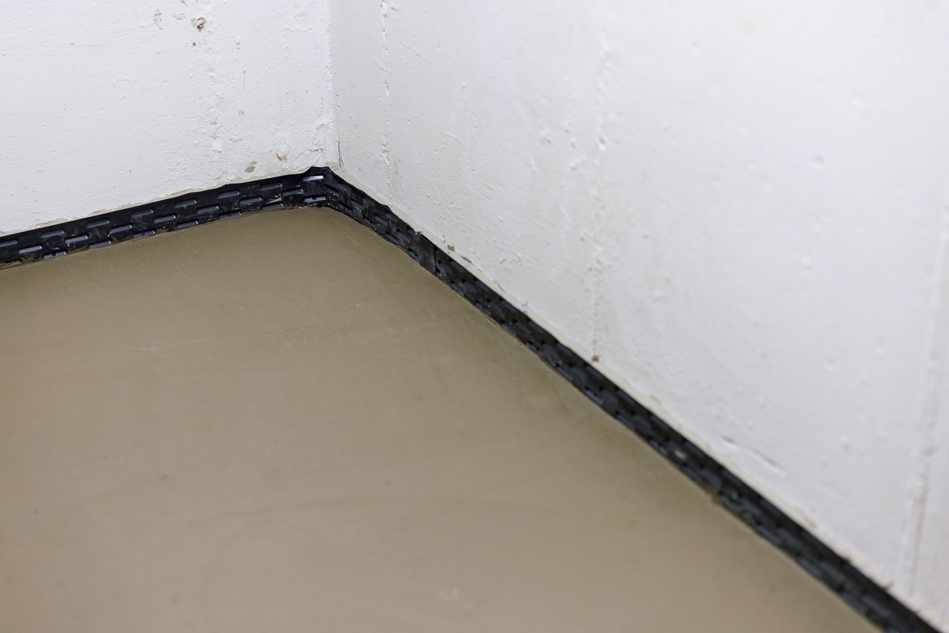 Concrete meets waterproof barrier in residential corner. Newly completed foundation moisture control system features fresh floor pour against dark protective membrane