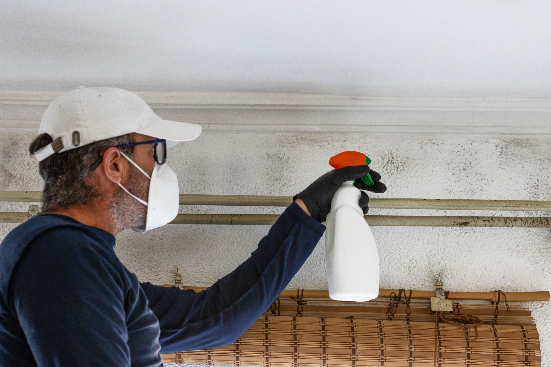 A man wearing a cap and a mask cleans mould from walls and ceilings with a sprayer.