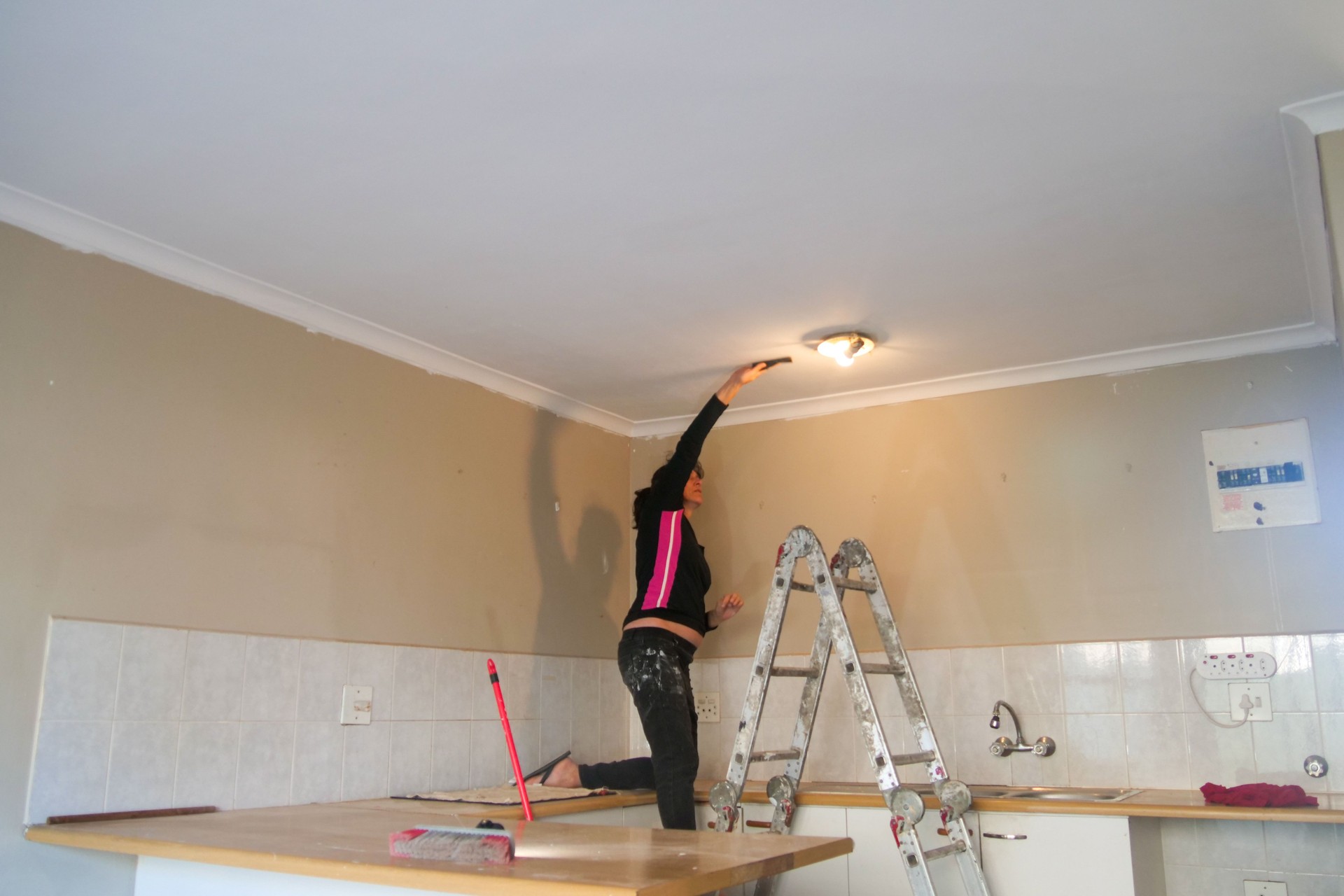 Woman standing on a kitchen countertop and sanding a ceiling while doing home improvements.